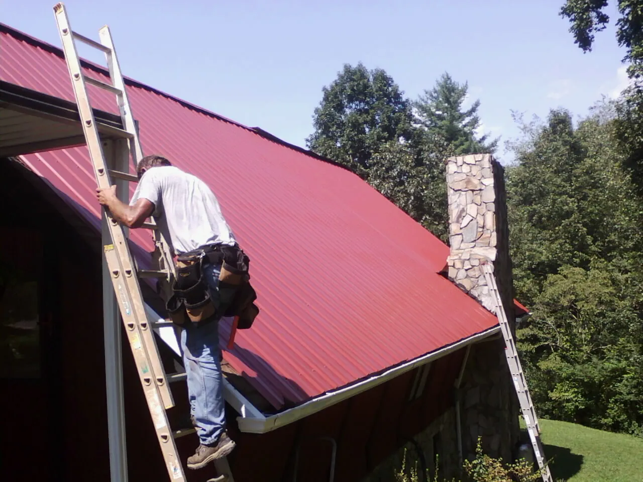 Roofer on a ladder beside a red metal roof and stone chimney — Soffit & Fascia Repair by Roofing Brothers in Mississippi State